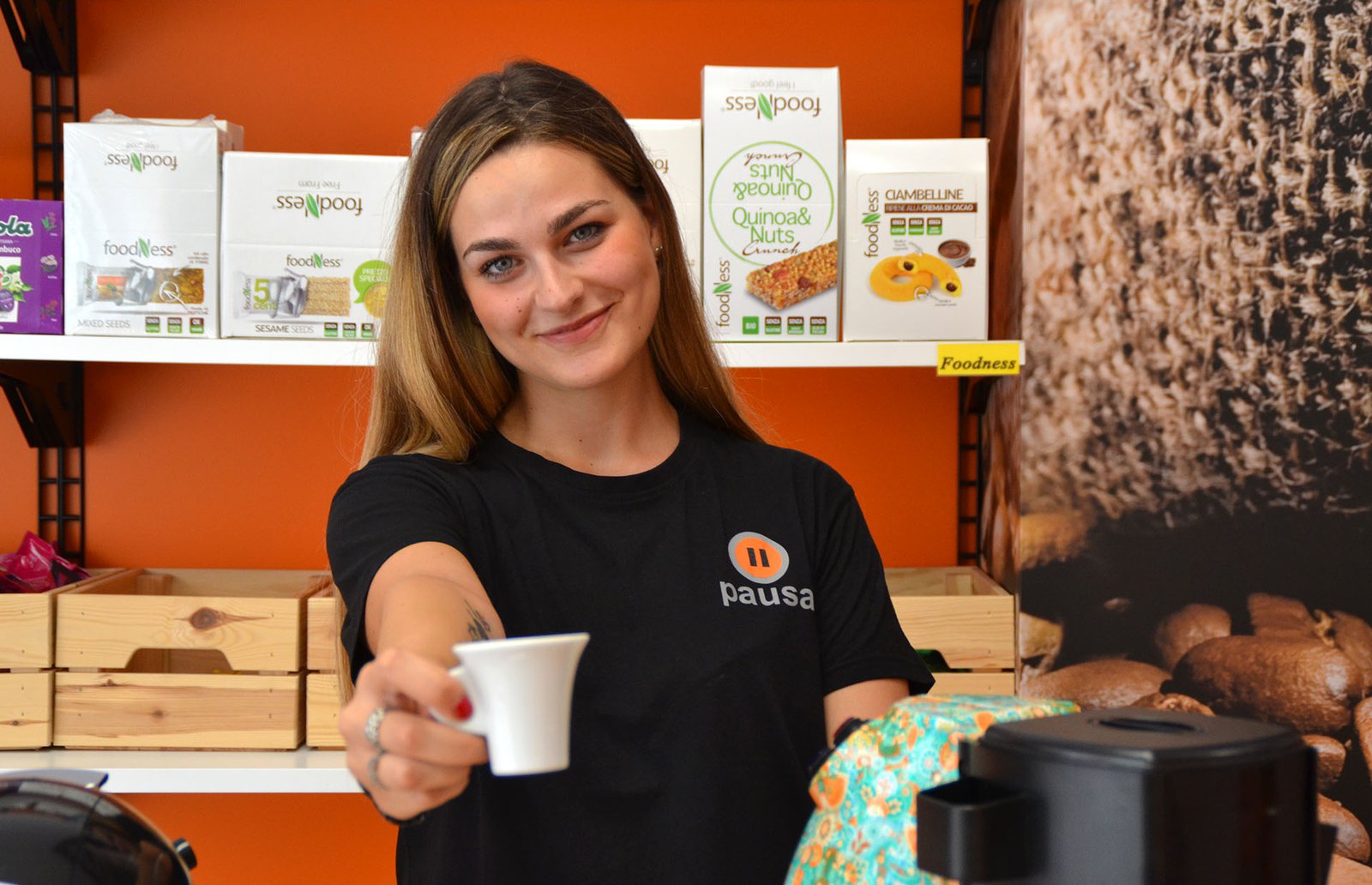 Ragazza con maglietta nera Pausa porge una tazzina bianca davanti a scaffali di snack Foodness su parete arancione